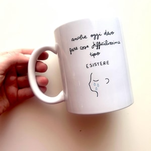 Può includere: Tazza in ceramica bianca con manico curvo. La tazza presenta testo scritto a mano e una semplice illustrazione di un volto che piange. Il testo sulla tazza è in italiano.