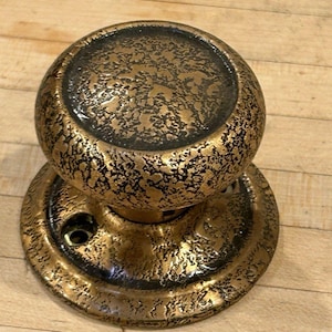 May include: Ornate, antique-style door knob with a textured, bronze-gold finish and black accents. The round knob sits on a matching circular base, showcasing a vintage aesthetic. Ideal for adding a touch of classic elegance to any door.