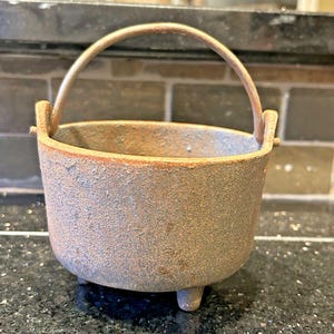 Pode incluir: Um pequeno caldeirão rústico de ferro fundido com uma alça curva. O caldeirão tem um corpo arredondado, três pés pequenos e uma superfície desgastada e texturizada com tons de castanho, azul e laranja. Ideal para fins decorativos ou culinária em pequena escala.