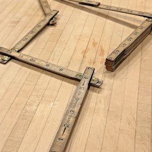 May include: Several antique wooden folding rulers are arranged on a light-colored wooden surface. The rulers are tan with black markings and brass hinges. One ruler is partially folded, displaying measurements up to 43 inches.