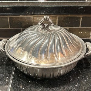 Könnte beinhalten: Silberfarbene Servierschüssel mit Deckel. Die Schüssel hat ein geripptes Design auf Deckel und Körper, mit dekorativen Griffen auf beiden Seiten. Der Deckel hat einen dekorativen Griff. Die Schüssel ist wahrscheinlich aus Metall.