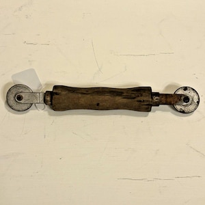 May include: An antique seam roller with a wooden handle and two metal wheels. The handle is dark brown and shows signs of wear. The wheels are circular and attached to the handle with metal supports. This tool is likely used for sewing or crafting.