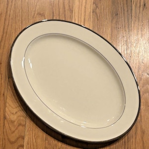 May include: An oval serving platter with a cream-colored surface and a thin, dark gray line near the edge. The rim is trimmed with a silver-colored band. The platter is on a wooden surface.