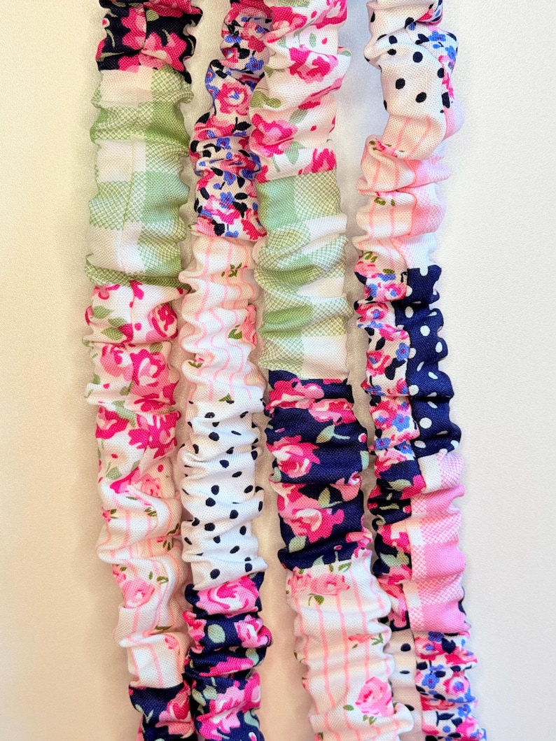 May include: Four long, fabric-covered elastic bands arranged vertically. Each band features a patchwork design of various patterns including pink roses, green gingham checks, navy blue polka dots, and thin pink stripes on a white background.