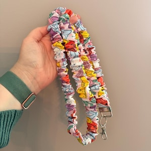Op de afbeelding: Een stoffen lanyard met een geplooid scrunchie-ontwerp en een kleurrijk patroon van schoolspullen, waaronder linialen, potloden en gummen. De band is afgewerkt met een zilverkleurige metalen draaisluiting en een bijpassende sleutelring.