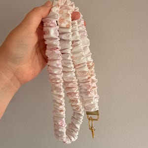 Op de afbeelding: Een stoffen lanyard met een geplooid, gerimpeld ontwerp in wit met een delicaat roze en oranje bloemenpatroon. De band is bevestigd aan een goudkleurige metalen sluiting en een kleine ronde ring, een functioneel accessoire voor sleutels of ID.