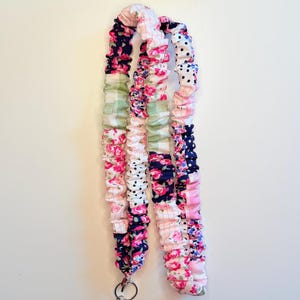 May include: A fabric lanyard featuring a gathered, scrunchie-style design with a patchwork pattern of floral prints, navy blue polka dots, and light green gingham checks. The strap includes a silver-toned metal swivel lobster clasp at the end.