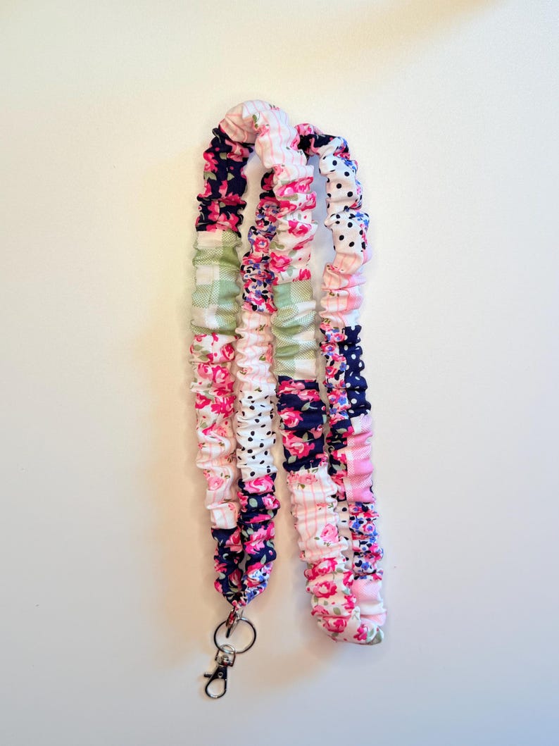 May include: A fabric lanyard featuring a gathered, scrunchie-style design with a patchwork pattern of floral prints, navy blue polka dots, and light green gingham checks. The strap includes a silver-toned metal swivel lobster clasp at the end.