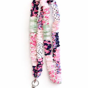 May include: A fabric lanyard with a variety of floral and patterned designs. The lanyard features a mix of pink, blue, and green fabrics with floral, polka dot, and gingham patterns. It has a silver-colored clasp.