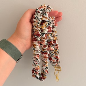 Op de afbeelding: Een stoffen lanyard met een geplooid, gerimpeld ontwerp op een witte achtergrond met een bloemenpatroon in tinten rood, oranje, geel en groen. De band is voorzien van een goudkleurige metalen draaibare karabijnhaak en een bijpassende ring.