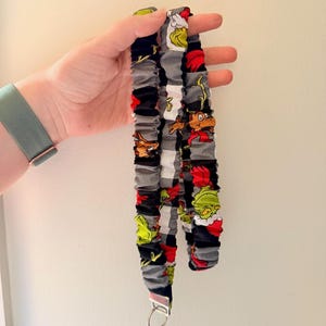 May include: A fabric lanyard featuring a repeating pattern of green characters in red hats and brown reindeer on a black, white, and gray buffalo plaid background. The lanyard is gathered with elastic and includes a silver metal swivel lobster clasp.