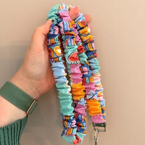 May include: A colorful lanyard with a metal clasp. The lanyard is made of fabric with a variety of patterns, including stripes, polka dots, and geometric shapes in shades of blue, pink, orange, and green. The lanyard is held in a hand.