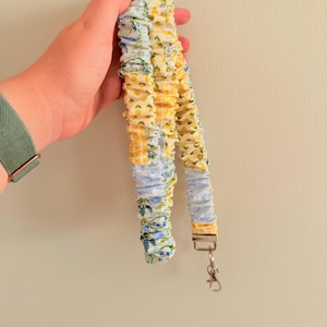 Op de afbeelding: Een bloemenlanyard met een metalen sluiting. De lanyard is gemaakt van stof met blauwe, gele en witte bloempatronen. De lanyard wordt tegen een lichtgroene muur gehouden. Ongeveer 60 cm lang.