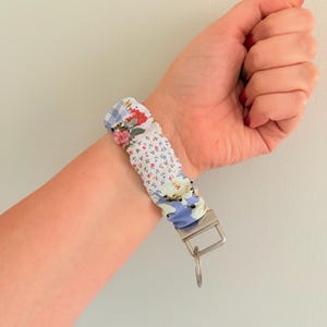 Puede incluir: Un llavero de pulsera de tela con un diseño de patchwork que incluye cuadros vichy azules, patrones de flores pequeñas y estampados de rosas rojas y blancas. La tela está fruncida sobre una banda elástica y unida a un cierre metálico plateado.
