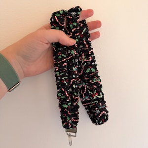 May include: A black fabric lanyard with a Christmas candy cane and holly pattern. The lanyard has a metal clasp and key ring. The fabric is gathered, creating a textured appearance. The background is a plain white wall.