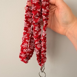 May include: A red fabric lanyard featuring a repeating white snowflake pattern. The lanyard is gathered with an elasticized design and includes a silver-toned metal swivel clasp and a circular split ring at the bottom for attaching items.