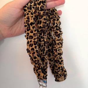Könnte beinhalten: Ein Stoff-Lanyard mit einem Leopardenmuster in Braun, Beige und Schwarz. Das Band hat eine geraffte, elastische Textur und ist mit einem silberfarbenen Metall-Karabiner sowie einem Schlüsselring am unteren Ende zur sicheren Befestigung versehen.