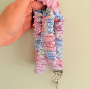 May include: A fabric lanyard featuring a gathered, scrunchie-style design with a patchwork pattern of light blue gingham, pink floral prints, and solid pink polka dots. The lanyard includes a silver-toned metal clasp and a circular key ring.