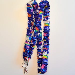 May include: A blue lanyard with a repeating pattern of unicorns, rainbows, and stars. The lanyard has a silver-colored metal clasp and a red-colored lobster claw clasp. The fabric is gathered, creating a textured appearance.
