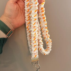 Op de afbeelding: Een stoffen lanyard met een herhalend patroon van gele potloden met roze gummen op een witte achtergrond. De lanyard is geplooid in een scrunchie-stijl en is voorzien van een zilverkleurige metalen sluiting en een ring aan de onderkant.