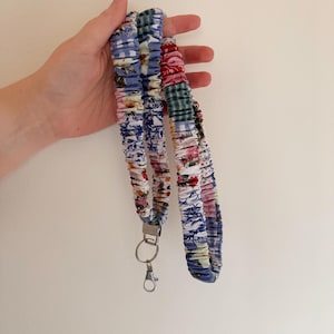 Może przedstawiać: Smycz z tkaniny z różnymi kwiatowymi i wzorzystymi wzorami w kolorze niebieskim, czerwonym i zielonym. Smycz ma srebrny metalowy karabińczyk i kółko na klucze. Tkanina jest marszczona i zszywana, tworząc teksturowany wygląd.