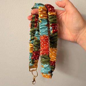 Op de afbeelding: Een stoffen lanyard met een geplooid scrunchie-ontwerp en een patchworkpatroon van bloemen- en geometrische prints in tinten blauwgroen, mosterdgeel, olijfgroen en donkerrood, afgewerkt met een goudkleurige metalen sluiting en sleutelring.
