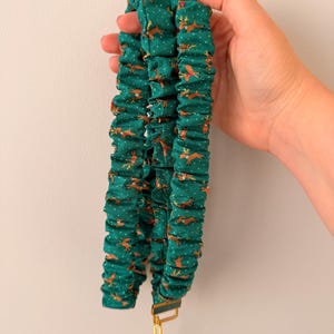 May include: A fabric lanyard featuring a dark green background with a repeating pattern of brown reindeer and small white dots. The lanyard is gathered in a ruffled style and includes a gold-toned metal swivel clasp at the bottom end.