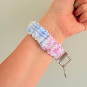 Puede incluir: Un llavero de pulsera de tela con una correa fruncida estilo coletero, que presenta un patrón de cuadros vichy azul y blanco en un lado y un estampado floral rosa en el otro, acabado con un cierre de metal plateado y una anilla circular de metal.