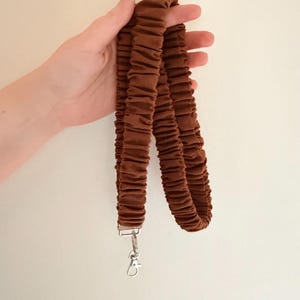 May include: A brown fabric lanyard featuring a gathered, scrunchie-style design. The strap is made of a solid brown material with a textured, ruffled appearance and is finished with a silver-toned metal swivel lobster clasp at the bottom end.