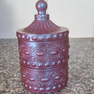 May include: A decorative, iridescent purple trinket box with a lid and a small spherical handle. The box has a detailed, embossed design with repeating patterns of circles and swirls. The color shifts from purple to blue.