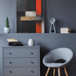 Puede incluir: Una pintura abstracta con formas rectangulares negras, rojas y blancas cuelga sobre una cómoda gris. Una silla gris con patas de madera está cerca. Una máquina de escribir vintage y un jarrón decorativo están en la cómoda.