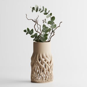 Könnte beinhalten: Beige Keramikvase mit einem wurzelartigen Design im unteren Bereich, die grüne Eukalyptusblätter und braune Zweige enthält. Die Vase hat eine zylindrische Form mit einer breiten Öffnung. Der Hintergrund ist schlicht weiß.