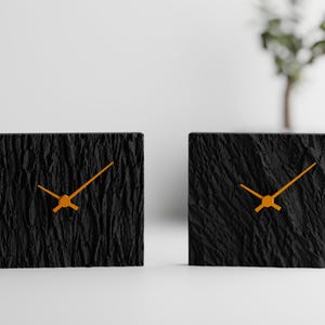 Könnte beinhalten: Zwei quadratische, schwarze Uhren mit strukturierten Oberflächen und orangefarbenen Zeigern. Die Uhren stehen nebeneinander auf einer weißen Oberfläche. Die Zeiger zeigen auf unterschiedliche Uhrzeiten. Im Hintergrund befindet sich eine dekorative Pflanze.