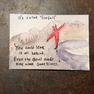 Könnte beinhalten: Eine Aquarellzeichnung auf Papier, die eine rote Teufelsfigur am Rande einer Klippe zeigt. Der Text lautet: It's called "freefall" You could leave it all behind, Even the devil needs time alone sometimes. Im Hintergrund sind blasse Berge zu sehen.