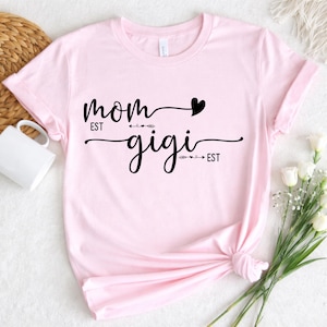Op de afbeelding: Lichtroze t-shirt met de woorden "mom" en "gigi" in zwart script. Een zwart hart staat boven het woord "mom". Het woord "est" staat boven en onder de woorden "mom" en "gigi".