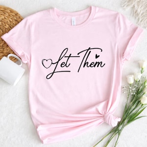 Op de afbeelding: Een lichtroze t-shirt met de zwarte tekst "Let Them". Een hartje staat voor het woord "Let" en een hartje volgt het woord "Them". Het t-shirt is aan de onderkant geknoopt.
