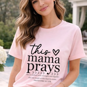 Op de afbeelding: Een lichtroze t-shirt met de tekst "this mama prays" in zwarte letters en blokletters. Het shirt bevat ook het bijbelvers Spreuken 31:25. Het shirt heeft korte mouwen en een ronde hals.