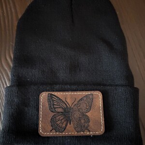 May include: A black beanie hat with a rectangular brown leather patch. The patch features a detailed, etched butterfly design. The hat is made of a knitted material, and the patch is stitched onto the folded brim.
