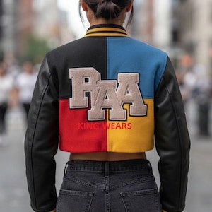 Pode incluir: Uma jaqueta varsity curta com mangas de couro preto e um painel traseiro em blocos de cores preto, azul, vermelho e amarelo. As costas exibem grandes letras de feltro bege formando PAA com o texto vermelho KING WEARS impresso abaixo.