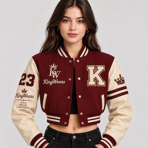 Op de afbeelding: Een kort varsity-jack in bordeauxrood met crèmekleurige mouwen. De voorkant is voorzien van een grote letter K, de tekst KingWears met een kroon en het getal 23. De mouwen bevatten kroonafbeeldingen en de tekst Style Builds Confidence.