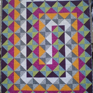 May include: A quilt with a geometric pattern of triangles in shades of orange, green, purple, and white. The quilt is bordered with a wide band of pink and orange.