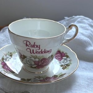 May include: A white porcelain teacup and saucer set featuring a gold-trimmed rim and handle. The cup displays the text Ruby Wedding in red script, accompanied by a floral pattern of red and white roses on both the cup and the saucer.