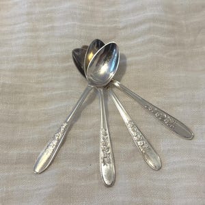 Könnte beinhalten: Ein Set aus vier silberfarbenen Metalllöffeln, die fächerförmig angeordnet sind. Jeder Löffel hat eine polierte ovale Laffe und einen schlanken Stiel, der mit einem geprägten Blumenmuster verziert ist und in einer glatten, abgerundeten Spitze endet.