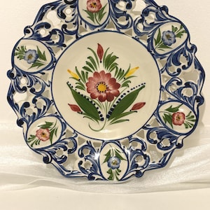 May include: A round ceramic plate featuring a central hand-painted floral motif in red, yellow, and green. The wide, scalloped border has intricate blue scrollwork and cut-out sections, interspersed with small oval panels containing painted flowers.