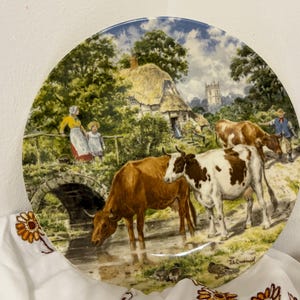 May include: A decorative plate featuring a pastoral scene with cows drinking from a stream. The plate depicts a bridge, a thatched-roof cottage, and figures in the background. The color palette includes greens, browns, and blues.