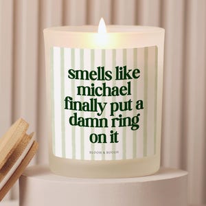 May include: A frosted glass candle with a light green and white vertical striped label featuring the dark green text: smells like michael finally put a damn ring on it. The brand name BLOOM & BOUGH is printed in small letters at the bottom.