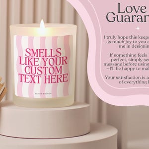 Puede incluir: Una vela encendida en un tarro de vidrio esmerilado con tapa de madera. La vela tiene una etiqueta ondulada rosa y blanca que dice "SMELLS LIKE YOUR CUSTOM TEXT HERE". La vela est&aacute; sobre un pedestal blanco.