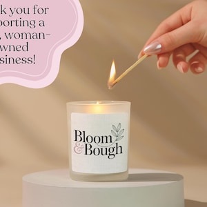 Puede incluir: Una vela encendida en un tarro de vidrio esmerilado con el texto "Bloom & Bough" en una etiqueta blanca. Se est&aacute; usando una cerilla para encender la vela. La imagen incluye el texto "&iexcl;Gracias por apoyar a una peque&ntilde;a empresa propiedad de mujeres!"