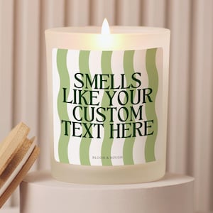 Puede incluir: Una vela de vidrio esmerilado encendida con una etiqueta a rayas verdes y blancas que dice "Smells Like Your Custom Text Here". Una tapa de madera descansa junto a la vela. La vela est&aacute; sobre un pedestal blanco.