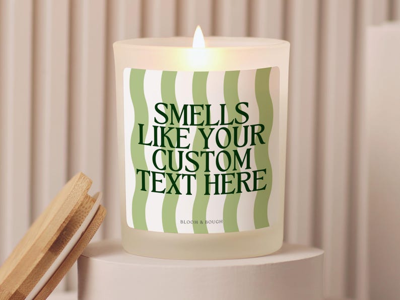 Puede incluir: Una vela de vidrio esmerilado encendida con una etiqueta a rayas verdes y blancas que dice "Smells Like Your Custom Text Here". Una tapa de madera descansa junto a la vela. La vela est&aacute; sobre un pedestal blanco.